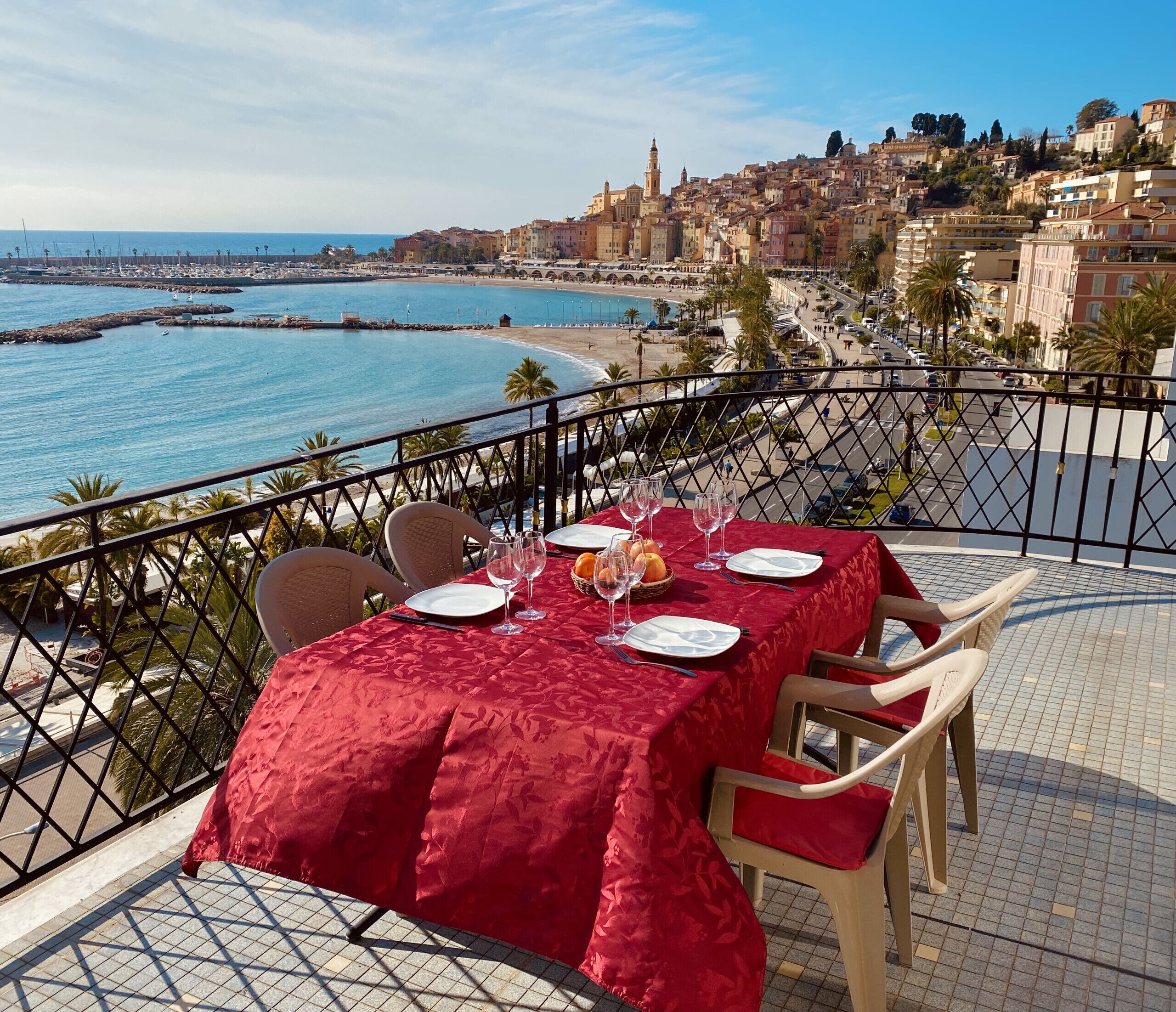 A table set for two with a red tablecloth, overlooking a coastal cityscape with a clear view of the sea and buildings.