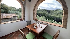 Un comedor con mesa y sillas, vista a un paisaje verde exuberante a través de ventanales arqueados, y una pared de piedra rústica.