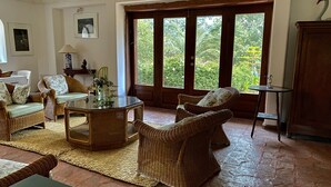 Un salon chaleureux, doté de meubles en osier, d’une table basse en verre et offrant une vue sur une végétation luxuriante grâce à de grandes fenêtres.