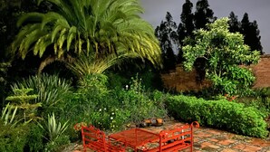 Un jardin avec un banc en métal rouge, diverses plantes et un mur de briques en arrière-plan.