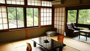 A traditional Japanese room with tatami flooring, a low table with a black tray, a cushioned bench, and a chair. Large windows with shoji screens provide views of greenery outside.