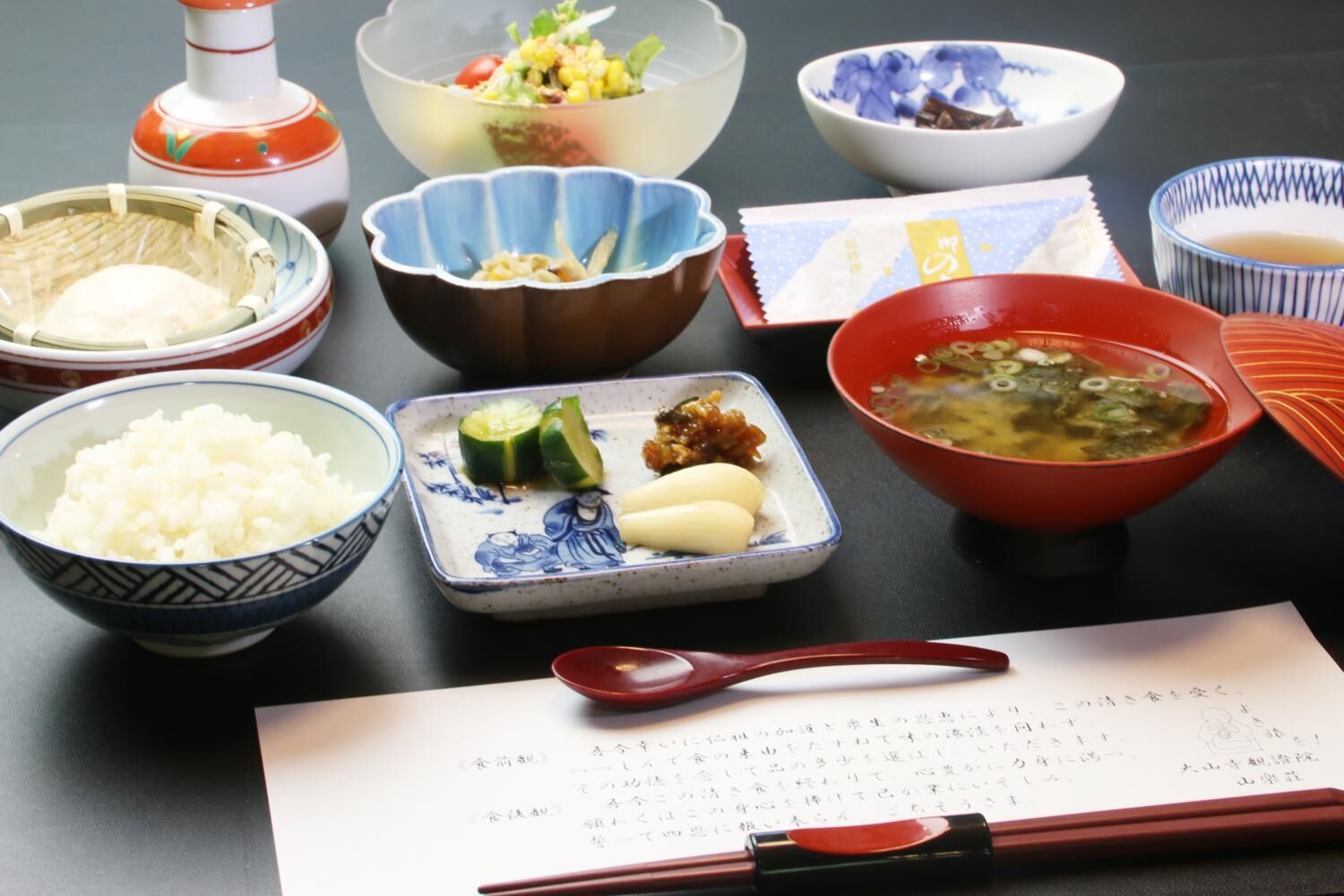 Une table où sont présentés divers plats japonais, notamment du riz, des légumes et une soupe, ainsi que des baguettes et un menu.
