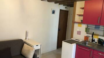 Un espace de vie compact comprenant un canapé, une petite table, un appareil électroménager blanc et une kitchenette dotée de meubles rouges.
