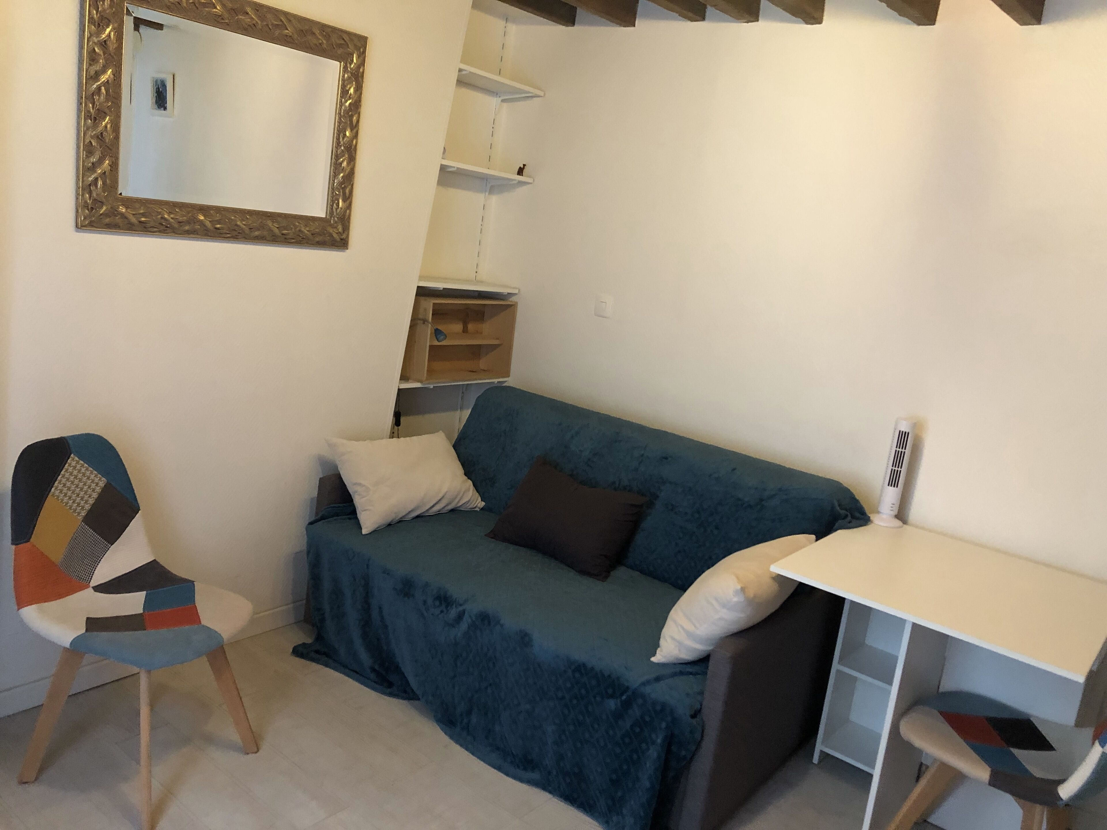 A small, cozy room with a blue sofa, a wooden chair, a white desk, and a mirror on the wall.