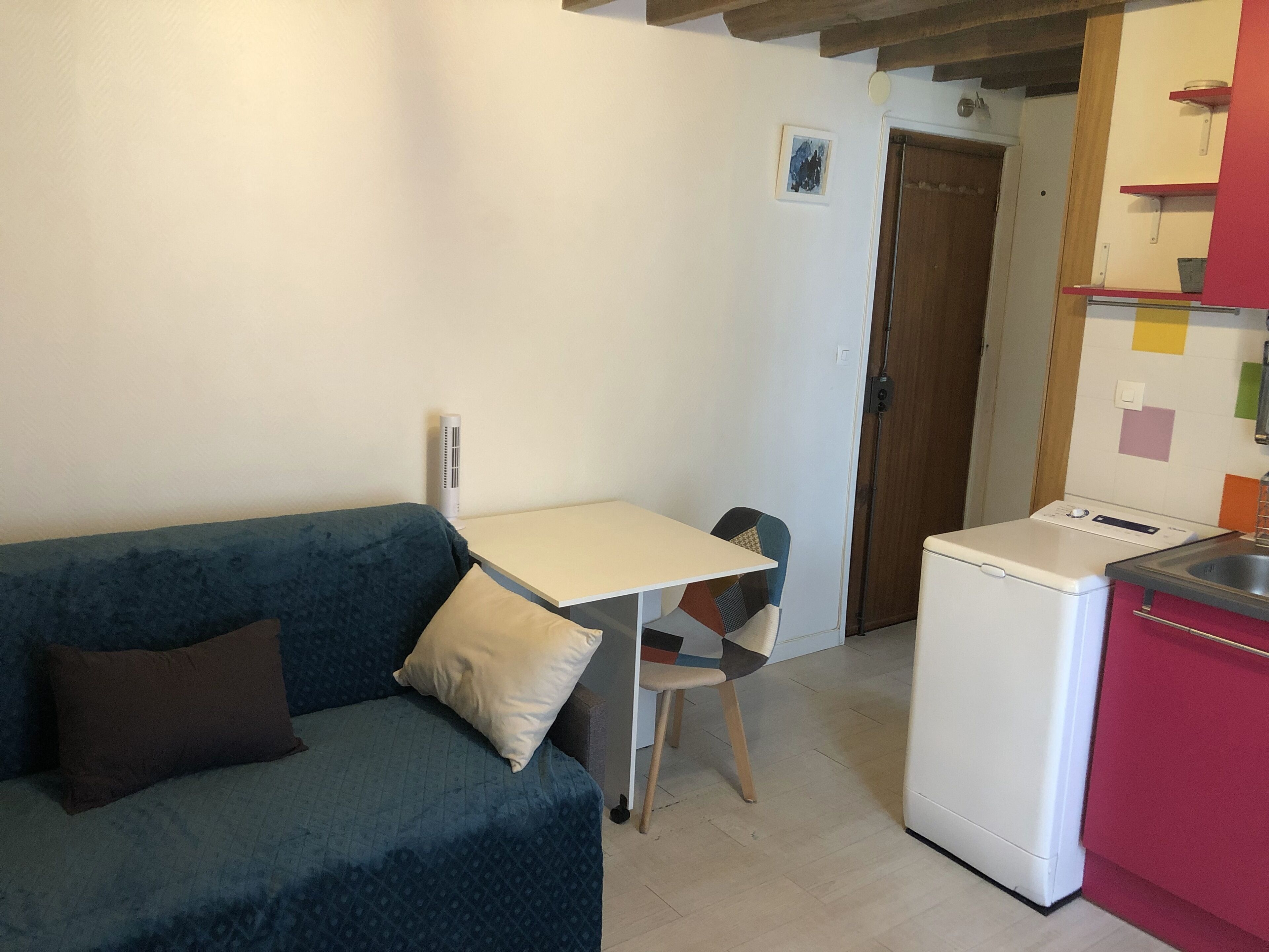 A small kitchenette with a white refrigerator, a small table, and a chair. There is a blue sofa with a pillow and a brown pillow.