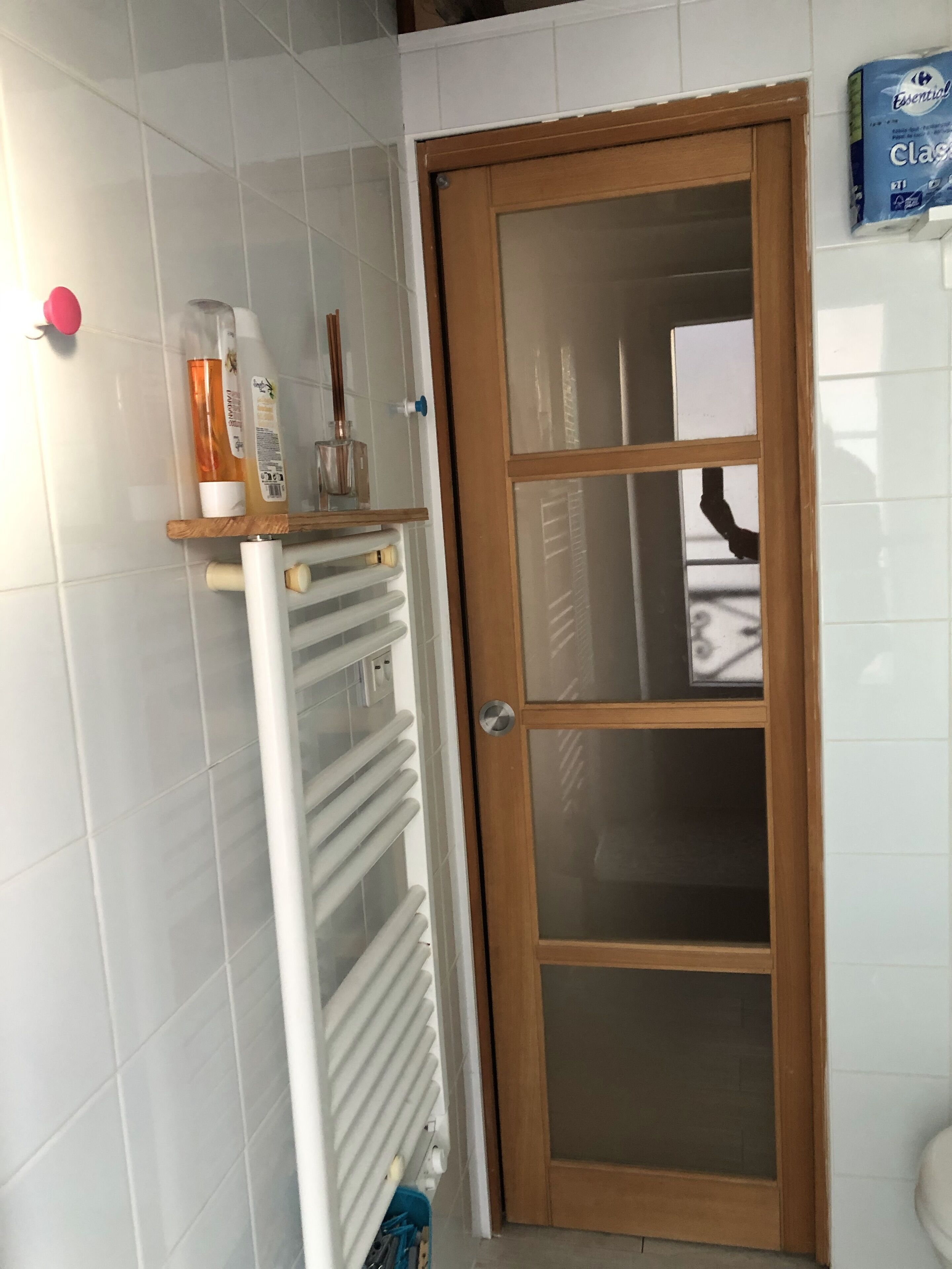 Une salle de bain avec un mur recouvert de carreaux blancs, une porte en bois avec des panneaux de verre, un radiateur et une étagère contenant des articles de toilette.