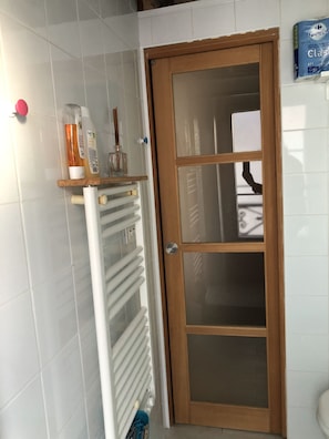 A bathroom with a white tiled wall, a wooden door with glass panels, a radiator, and a shelf with toiletries. - Beautiful studio, 1 or 2 people (Paris)