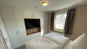A bedroom with a bed, a TV mounted on a cabinet, a window with curtains, and a radiator. - COMPLETELY RENOVATED! NO PRICE INCREASE FOR 2026+2027 !!! BEACH CHAIR INCLUDED! (Binz)