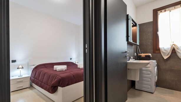 Ein Schlafzimmer mit einem Bett, einem Nachttisch und einem kleinen Tisch daneben. Es gibt ein Badezimmer mit Waschbecken, Spiegel und Handtuchhalter.