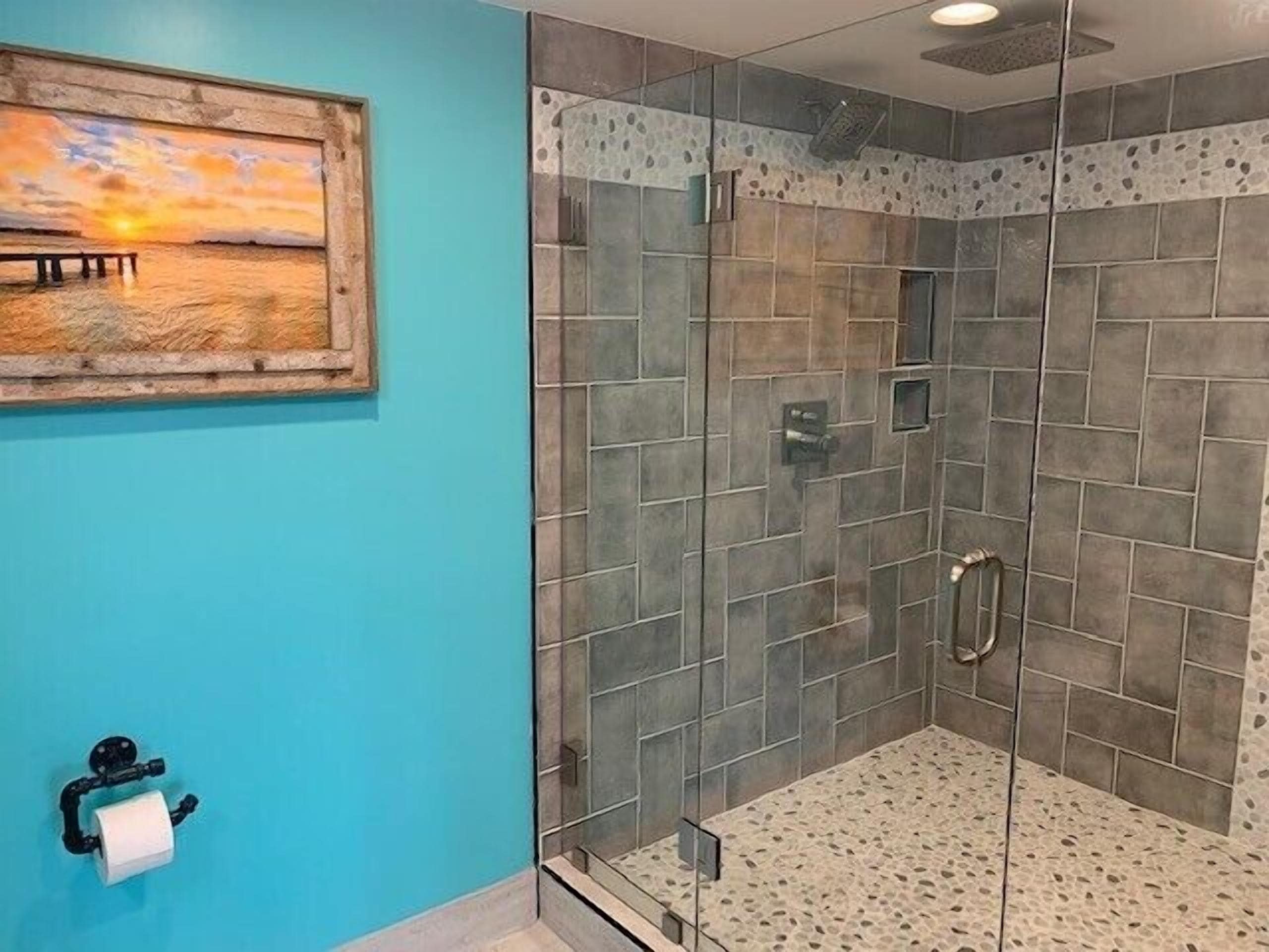 A bathroom with a glass shower enclosure, a toilet paper holder, and a painting of a sunset over water.