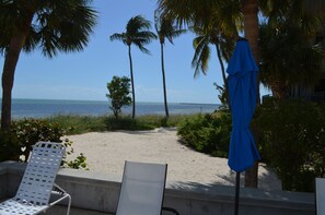 A beach with a white lounge chair and a blue umbrella, palm trees, and a clear view of the ocean.