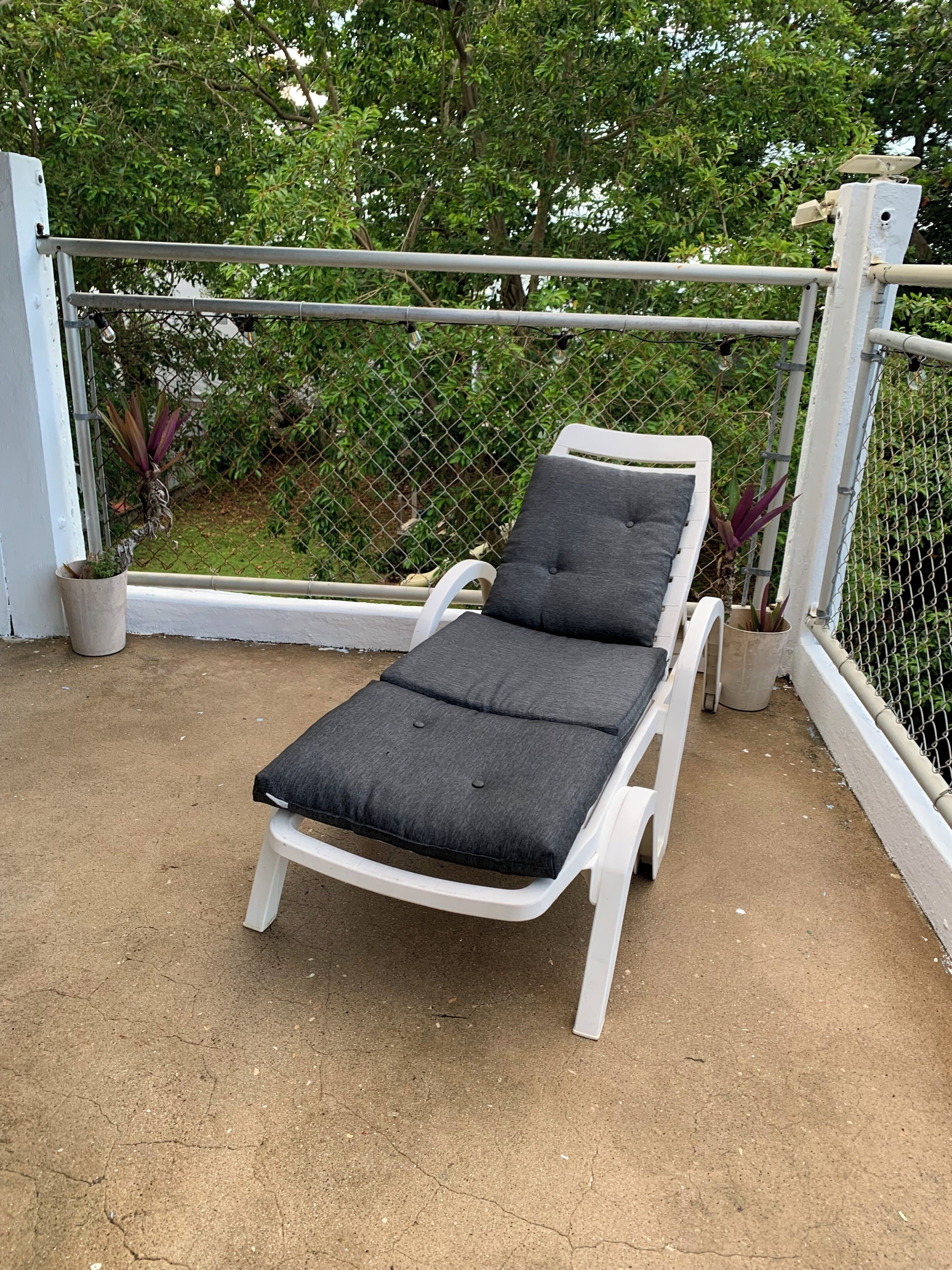 Silla de exterior blanca con un cojín negro, ubicada sobre una superficie de concreto, rodeada por una cerca de malla y follaje.