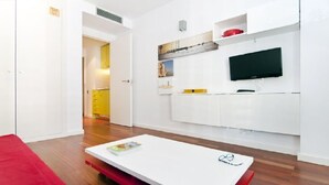 A modern, minimalistic living space with a flat-screen TV mounted on the wall, a small dining table, and a red sofa.