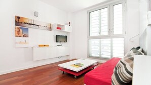 A modern living room with a white sofa, a red ottoman, a flat-screen TV, a white entertainment unit with artwork, and a large window.