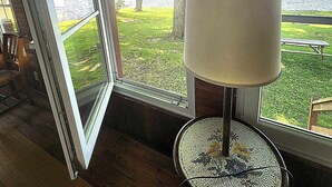 Une pièce dotée d’une grande fenêtre, d’une table ronde avec une lampe, et offrant une vue sur un espace verdoyant et un plan d’eau.