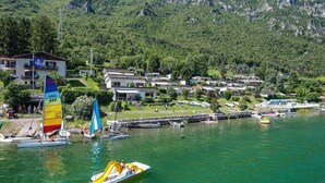 A lakeside view with a sailboat, a speedboat, and a dock with boats. There are buildings in the background and a mountainous landscape. - Directly on the lake with fenced garden (Tre Capitelli)