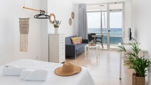 Et moderne hotelværelse med balkon, seng med hvide sengetøj, en grå sofa og et spiseområde med et bord og stole.