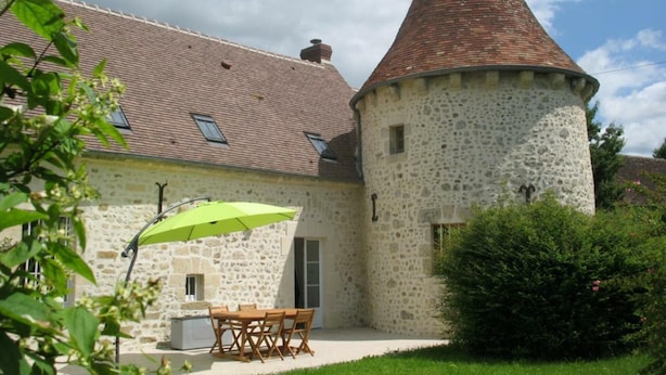 A stone building with a conical tower, a patio with a table and chairs, and a green umbrella. - Charming historic cottage, 5 bedrooms, large garden (Coulonges-sur-Sarthe)