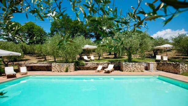 A swimming pool with lounge chairs and umbrellas, surrounded by trees and stone walls. - Ginestra (Montefalco)