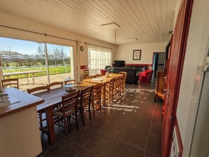 A dining area with a wooden table and chairs, a television, and a view of a green landscape through large windows.