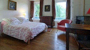 Un dormitorio con cama, escritorio, silla y cómoda. Hay dos ventanas con cortinas.