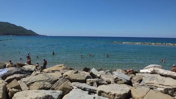 Une plage rocheuse bordée d’une eau d’un bleu limpide, avec un paysage vallonné en arrière-plan.