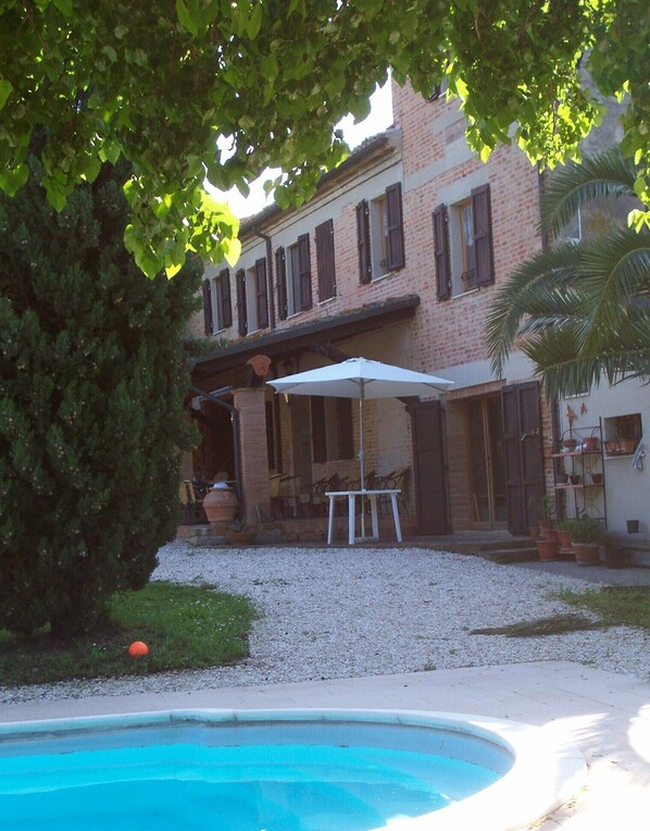 A pool area with a brick building in the background, surrounded by trees and gravel. - The Tower in Solebello (Morro D' Alba)