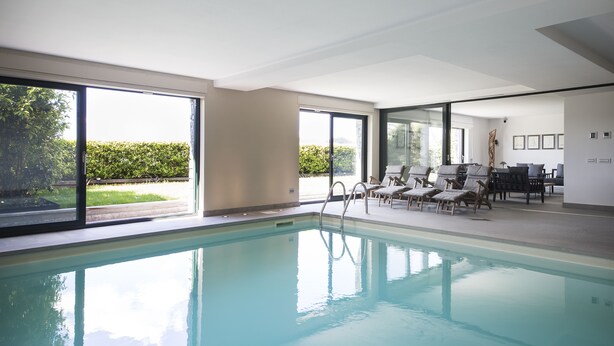 Une piscine intérieure avec des parois en verre transparent, un espace de détente au bord de la piscine et une vue sur l’extérieur.