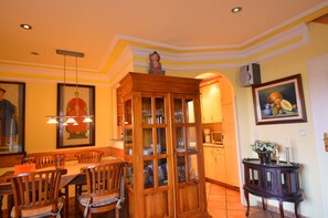 Een eetruimte met houten stoelen, een glazen kast met servies en een schilderij aan de muur.