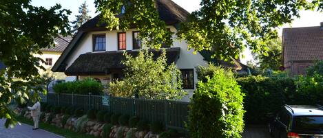 A traditional house with a thatched roof, surrounded by lush greenery and a stone wall.