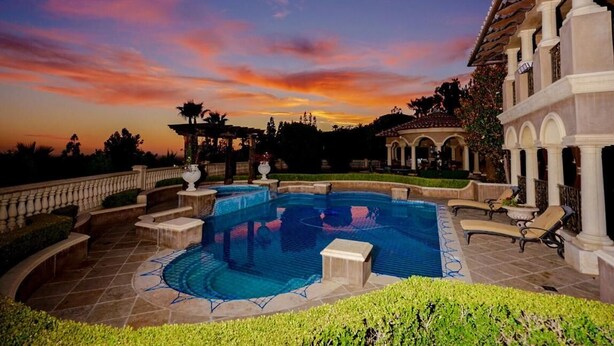 A large outdoor swimming pool surrounded by a landscaped garden, with a building featuring classical architecture in the background. - CASA VALENTINA-Beautiful Villa Estate (Whittier)