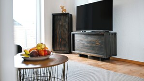 Ein modernes Wohnzimmer mit einem Flachbildfernseher auf einer Holzkassette, einem kleinen runden Tisch mit Früchten und einem großen Fenster, das natürliches Licht hereinlässt.