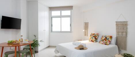 A modern, well-lit bedroom with a large bed, a small dining table, and a TV mounted on the wall.