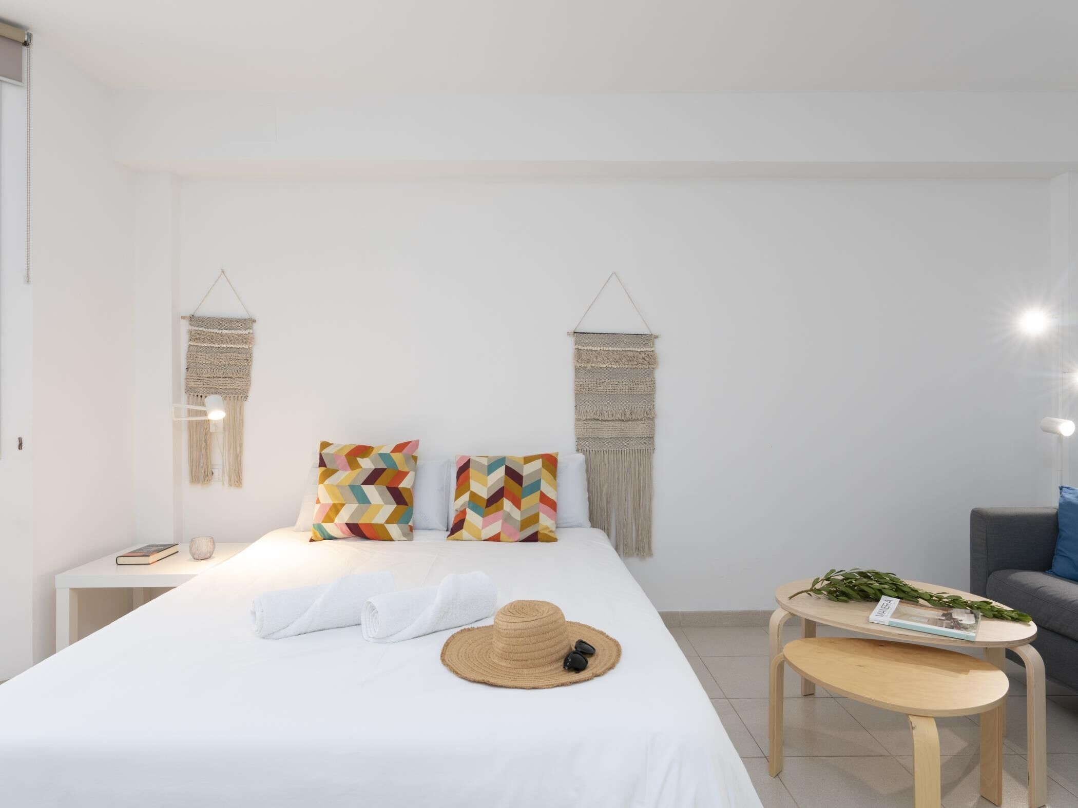 A minimalist hotel room with a bed, two macramé wall hangings, a small table with a plant, and a grey sofa.