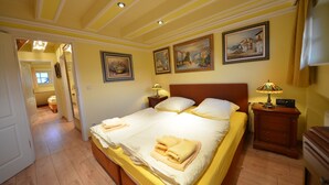 Ein Hotelzimmer mit einem großen Bett, zwei Nachttischen und einem Beistelltisch mit einer Lampe. An der Wand hängen drei Bilder und eine Tür führt in einen weiteren Raum.