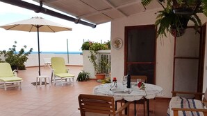 A terrace with a table set for two, surrounded by chairs, a potted plant, and a view of the sea.