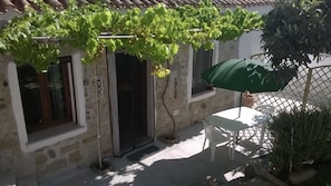 Un bâtiment en pierre surmonté d’une pergola couverte de plantes grimpantes vertes, une table et des chaises blanches, et un parasol vert.