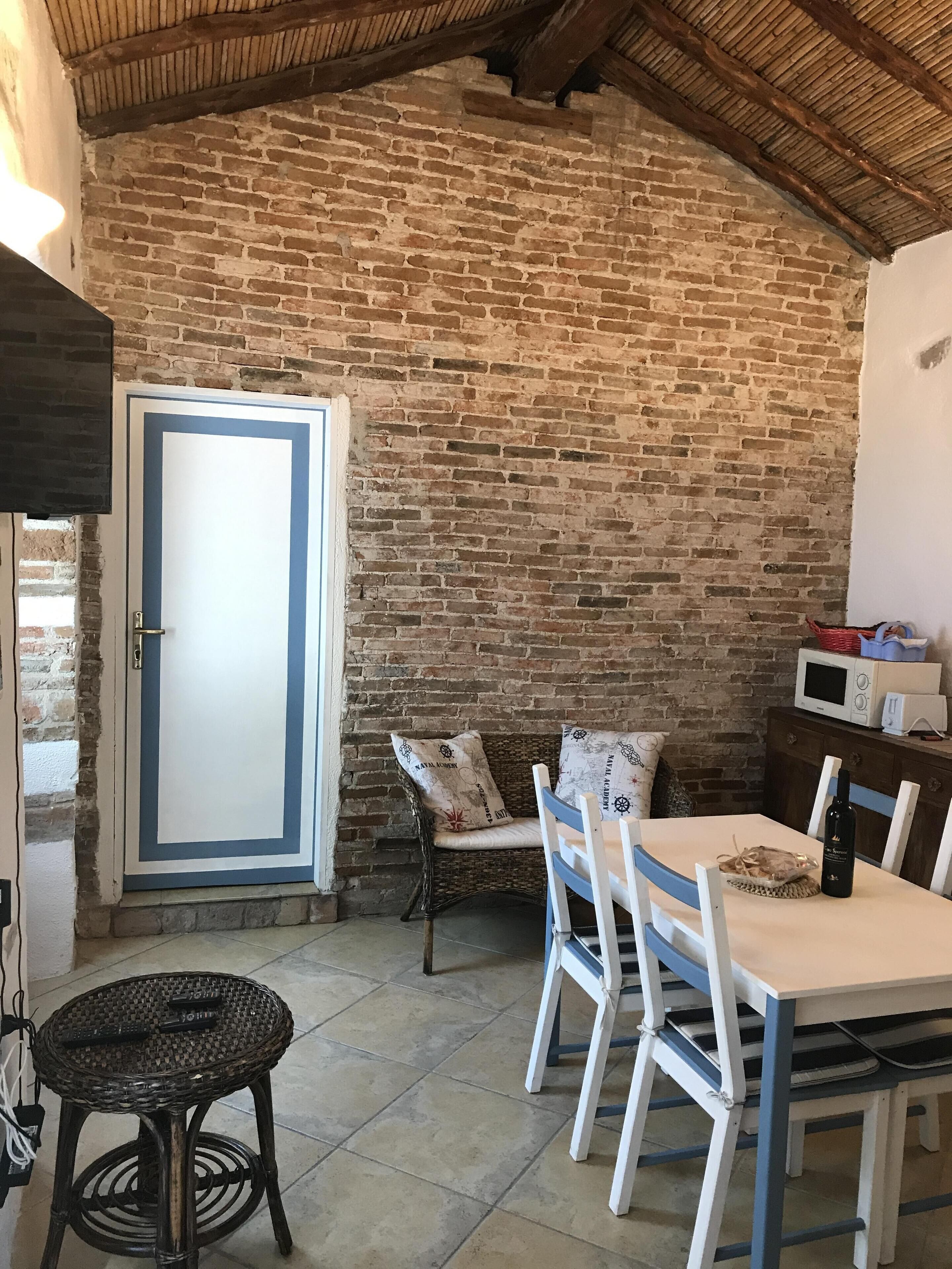 Una habitación rústica con pared de ladrillo, puerta blanca con marco azul, mobiliario de mimbre y una mesa con sillas.