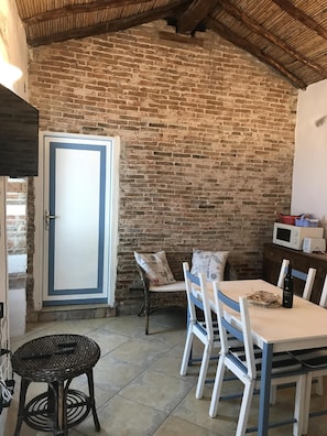 Une pièce rustique avec un mur en briques, une porte blanche dotée d’un cadre bleu, des meubles en osier et une table avec des chaises.