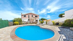 Una piscina con borde de piedra, una casa de dos pisos al fondo, y un cielo azul despejado.