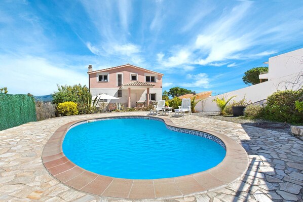A swimming pool with a stone border, a two-story house in the background, and a clear blue sky.