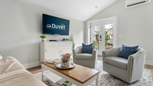 Un salon comprenant un canapé, deux fauteuils à rayures, une table basse ornée d’un vase décoratif, un téléviseur à écran plat fixé au mur et une vue sur l’extérieur, visible à travers une porte vitrée.