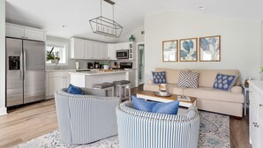 Une cuisine moderne avec des armoires blanches, des appareils en acier inoxydable et un îlot central. Le salon est agrémenté d’un canapé beige avec des coussins bleus et blancs, et un tapis à motifs recouvre le sol.