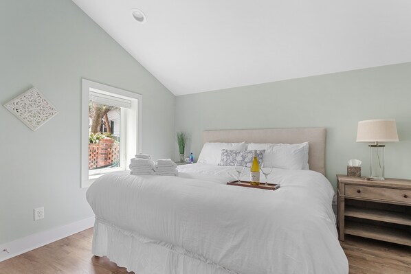 A bedroom with a large bed, a nightstand, a window with a view of a brick wall and plants, and a ceiling light.