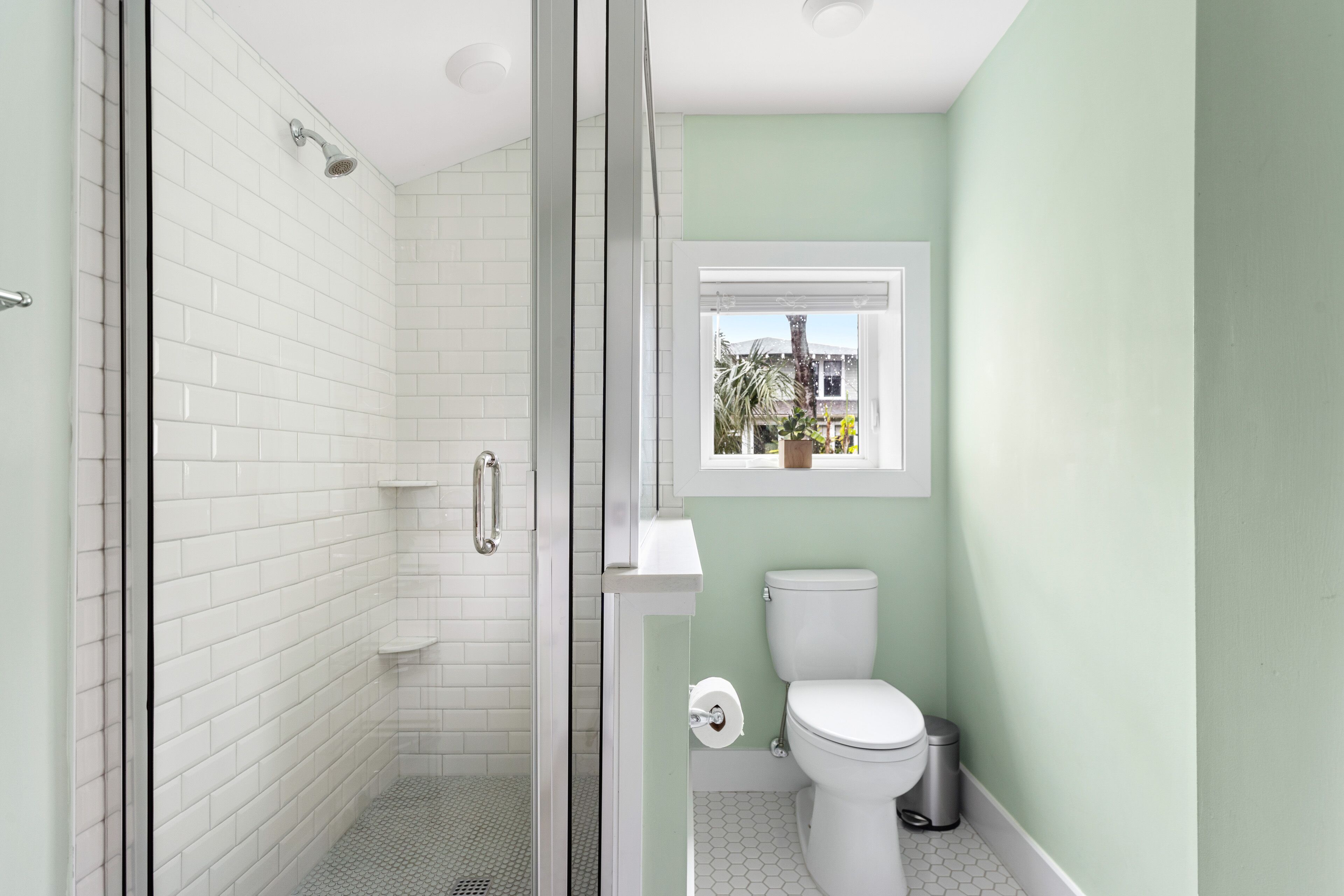 A bathroom with white subway tiles, a glass-enclosed shower, a toilet, and a window with a view of a house and trees.