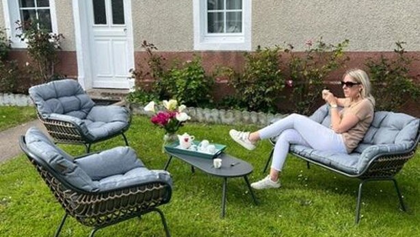 Un patio con dos sillas de mimbre, una mesita con un florero y una persona sentada en una de las sillas.