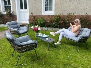 Une terrasse avec deux fauteuils en osier, une petite table sur laquelle est posé un vase de fleurs, et une personne assise sur l’un des fauteuils.