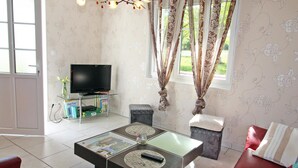 Una sala de estar con una mesa de centro de vidrio, un televisor sobre un mueble bajo, un sofá rojo y una ventana con cortinas florales.