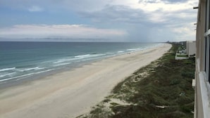 A beach view from a window overlooking a sandy beach with waves, a grassy area, and buildings in the distance. - Southpoint 707 La Casa Azul (Ponce Inlet)