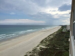 Vista a la playa desde una ventana, con arena, olas, una zona verde y edificios al fondo.